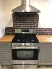 Kitchen with stainless steel range with gas stovetop, light tile flooring, wall chimney range hood, gray cabinets, and wood counters