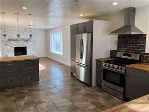 Kitchen featuring stainless steel appliances, wall chimney range hood, a fireplace, wood counters, and pendant lighting