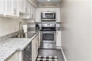 Kitchen featuring white cabinets, stainless steel appliances, sink, light stone counters, and dark tile flooring