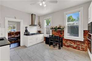 Kitchen featuring ceiling fan, light tile flooring, wall chimney range hood, tasteful backsplash, and white cabinetry