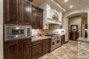 Kitchen featuring dark brown cabinets, appliances with stainless steel finishes, backsplash, light stone counters, and light tile floors