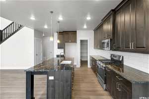 Kitchen featuring sink, light wood-type flooring, stainless steel appliances, dark stone countertops, and a kitchen island with sink