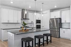 Kitchen with appliances with stainless steel finishes, light hardwood / wood-style floors, tasteful backsplash, wall chimney exhaust hood, and a kitchen island with sink