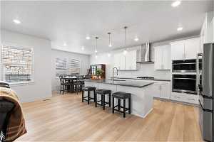 Kitchen with appliances with stainless steel finishes, a center island with sink, light hardwood / wood-style floors, and wall chimney exhaust hood