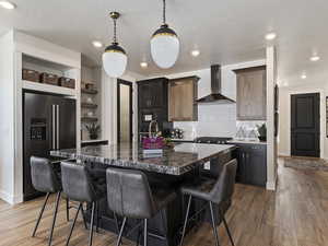 Kitchen featuring wood-type flooring, black appliances, backsplash, wall chimney range hood, and an island with sink