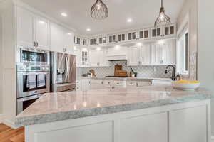 Kitchen featuring custom exhaust hood, tasteful backsplash, white cabinetry, hanging light fixtures, and stainless steel appliances