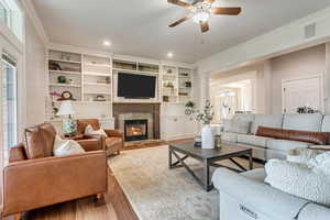 Living room with ceiling fan, built in shelves, a tiled fireplace, ornamental molding, and wood-type flooring