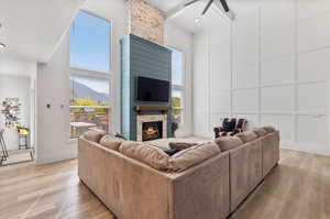 Living room with light hardwood / wood-style flooring, ceiling fan, a stone fireplace, beamed ceiling, and a high ceiling