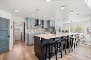 Kitchen featuring wall chimney range hood, light wood-type flooring, a center island with sink, backsplash, and pendant lighting