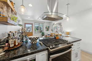 Kitchen with light wood-type flooring, dark stone counters, stainless steel range with gas stovetop, island exhaust hood, and white cabinets