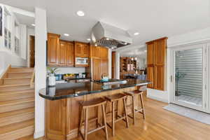 Kitchen featuring dark stone counters, light hardwood / wood-style flooring, island range hood, appliances with stainless steel finishes, and a breakfast bar area