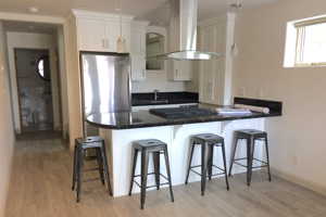 Kitchen with wall chimney range hood, white cabinetry, kitchen peninsula, and light hardwood / wood-style flooring