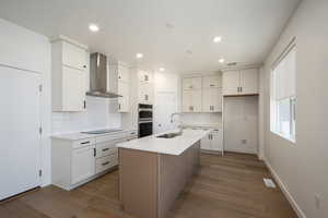 Kitchen with black electric stovetop, wall chimney exhaust hood, hardwood / wood-style flooring, sink, and a center island with sink