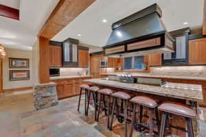 Kitchen with light tile flooring, premium range hood, stainless steel microwave, light stone counters, and backsplash