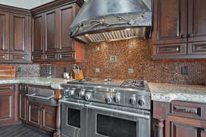 Kitchen with wall chimney range hood, backsplash, range with two ovens, and light stone counters