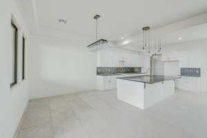 Kitchen featuring backsplash, hanging light fixtures, white cabinetry, an island with sink, and light tile floors