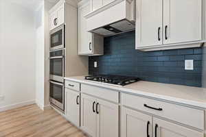 Kitchen featuring custom exhaust hood, stainless steel appliances, light wood-type flooring, backsplash, and white cabinets