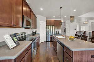 Kitchen with dark wood-type flooring, appliances with stainless steel finishes, a kitchen island with sink, pendant lighting, and sink