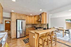 Kitchen with wall chimney range hood, light tile flooring, stainless steel appliances, backsplash, and a breakfast bar area