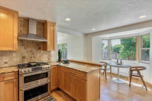 Kitchen with wall chimney range hood, kitchen peninsula, backsplash, double oven range, and light tile floors