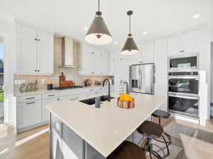 Kitchen with backsplash, wall chimney exhaust hood, an island with sink, light hardwood / wood-style floors, and stainless steel appliances