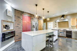 Kitchen featuring a center island, wall chimney range hood, hanging light fixtures, brick wall, and appliances with stainless steel finishes