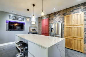 Kitchen with a center island, light stone countertops, brick wall, stainless steel fridge, and decorative light fixtures