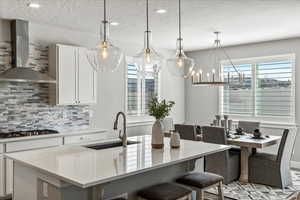 Kitchen featuring an island with sink, stainless steel gas stovetop, decorative light fixtures, backsplash, and wall chimney exhaust hood