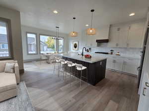 Kitchen with light wood-type flooring, an island with sink, hanging light fixtures, white cabinetry, and a breakfast bar