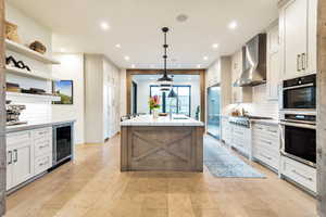 Kitchen featuring decorative light fixtures, a center island with sink, tasteful backsplash, wine cooler, and wall chimney exhaust hood