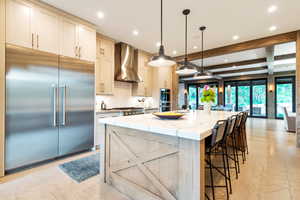 Kitchen with appliances with stainless steel finishes, beamed ceiling, wall chimney exhaust hood, pendant lighting, and a large island with sink