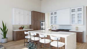 Kitchen featuring light hardwood / wood-style floors, white cabinetry, an island with sink, stainless steel double oven, and a high ceiling