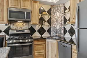 Kitchen with dark stone countertops, stainless steel appliances, backsplash, sink, and light brown cabinetry