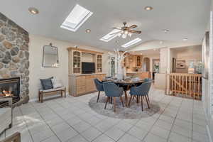 Dining area featuring ceiling fan, a fireplace, light tile flooring, and lofted ceiling with skylight