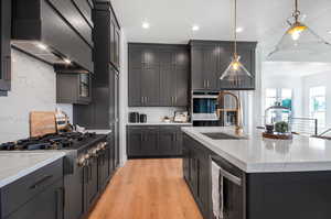 Kitchen with wall chimney range hood, light stone countertops, a center island with sink, and light hardwood / wood-style flooring