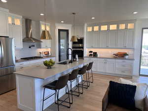 Kitchen with stainless steel appliances, wall chimney range hood, sink, light wood-type flooring, and an island with sink