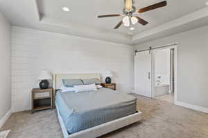 Carpeted bedroom featuring a barn door, ceiling fan, a tray ceiling, and ensuite bath