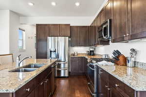 Kitchen featuring light stone counters, dark hardwood / wood-style floors, stainless steel appliances, a kitchen island with sink, and sink