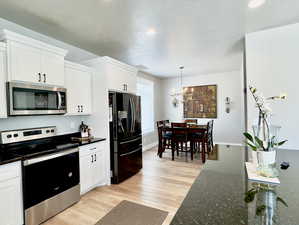 Kitchen featuring light wood-type flooring, dark stone countertops, a notable chandelier, white cabinets, and appliances with stainless steel finishes