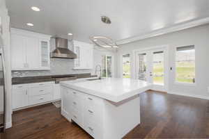 Kitchen featuring decorative light fixtures, wall chimney range hood, backsplash, a kitchen island with sink, and dark wood-type flooring