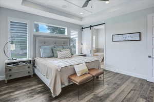 Bedroom featuring dark hardwood / wood-style flooring, ceiling fan, a raised ceiling, a barn door, and ensuite bath