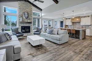 Living room featuring a stone fireplace, beamed ceiling, wood-type flooring, ceiling fan with notable chandelier, and high vaulted ceiling