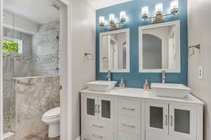 Primary bathroom featuring dual bowl vanity, marble tile shower, and pony wall toilet.