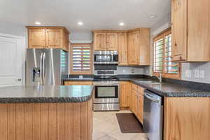 Kitchen center island, sink, light tile flooring, plantation shutters, durable stone countertops, and stainless steel appliances.