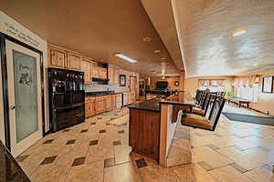 Kitchen with a breakfast bar area, light tile flooring, black fridge, a textured ceiling, and a kitchen island with sink.