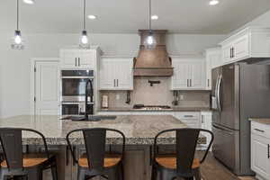 Kitchen with hanging light fixtures, appliances with stainless steel finishes, an island with sink, pot filler and premium range hood