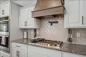 Kitchen with white cabinetry, stone countertops, custom range hood, double ovens, pot filler, stainless steel appliances, and backsplash