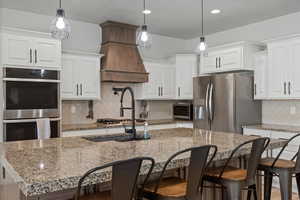 Kitchen featuring tasteful backsplash, a center island with sink, custom range hood, pot filler, white cabinetry, and appliances with stainless steel finishes