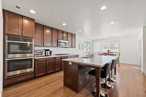 Kitchen with dark brown cabinets, light hardwood / wood-style floors, sink, a kitchen island with sink, and appliances with stainless steel finishes