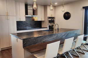 Interior space featuring wall chimney range hood, dark wood-type flooring, stainless steel double oven, a kitchen island with sink, and backsplash